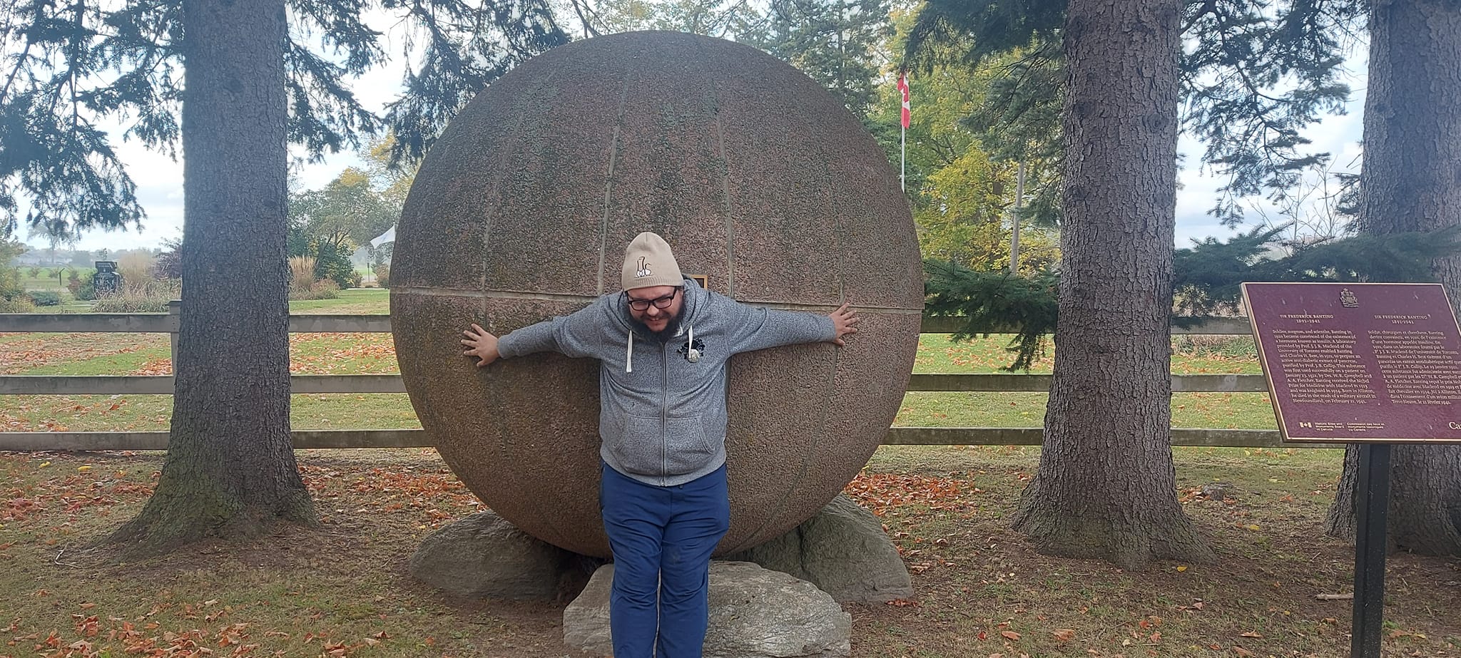 A man with short hair and bushy facial hair pretends to struggle to hold back a large concrete sphere in front of a wooden farm fence